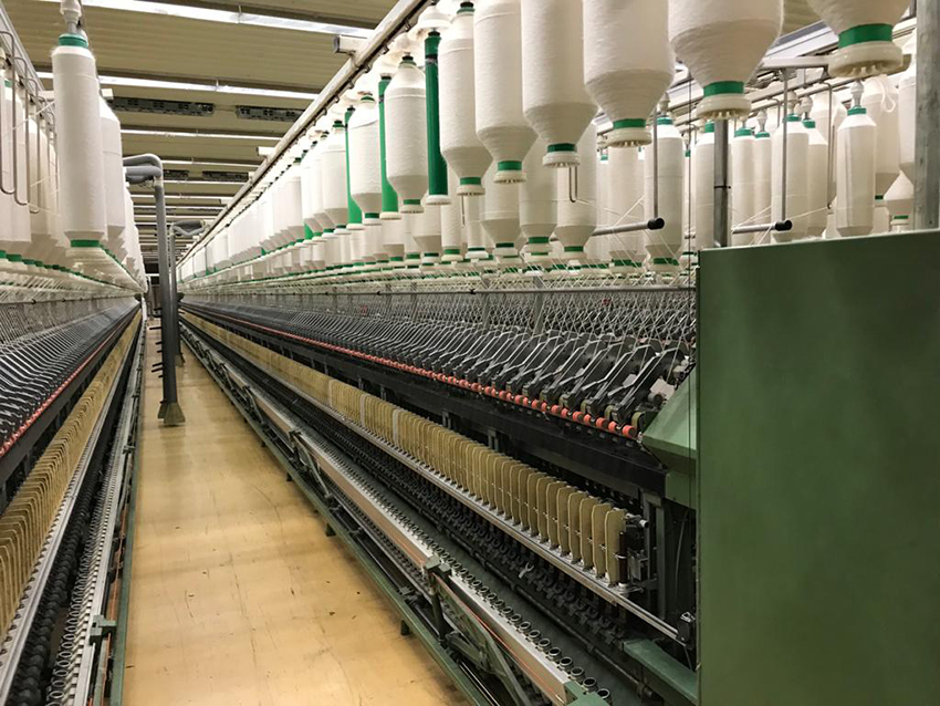 Een lange hal waar textielgaren op klossen aan het plafond hangen.