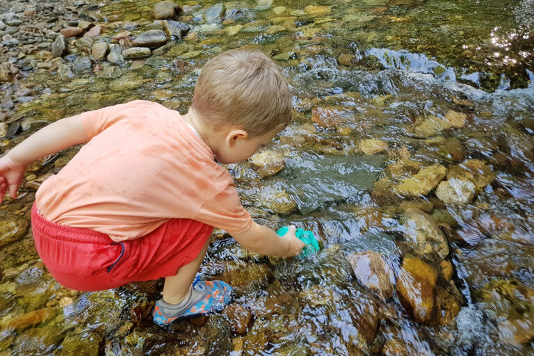 Der kleine Junge beugt sich über den flachen Bach und hält eine grüne Wasserbombe ins Wasser.