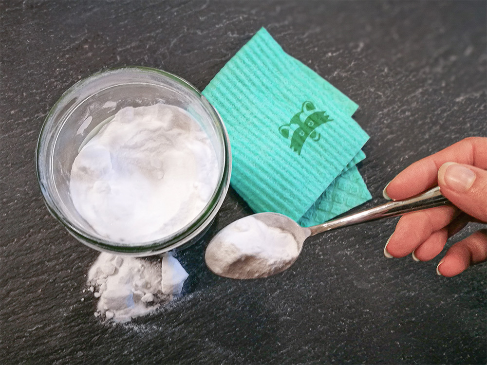 Eén hand houdt een lepel met natron/baking soda vast, daarnaast een glas en een sponsdoek