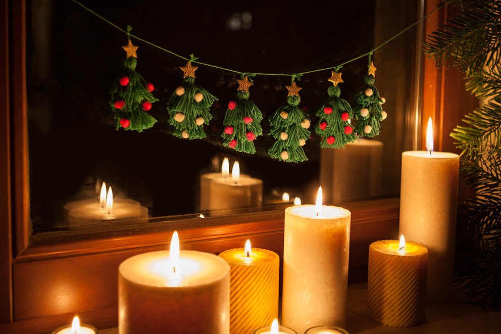 Die Girlande mit Weihnachtsbäumen aus Garn hängt an einem Fenster, auf dessen Fensterbank viele Kerzen stehen.