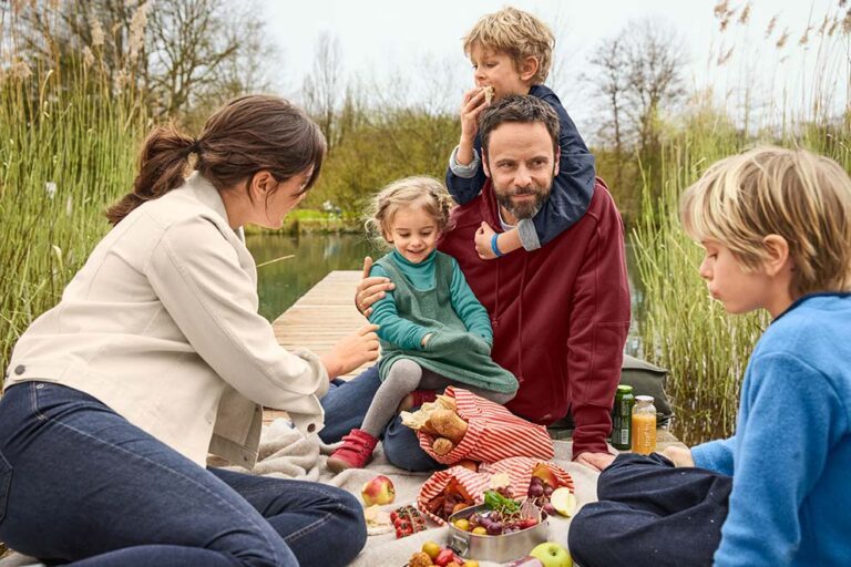 Eine Familie sitzt beim gemeinsamen Picknick auf einem Steg am See.