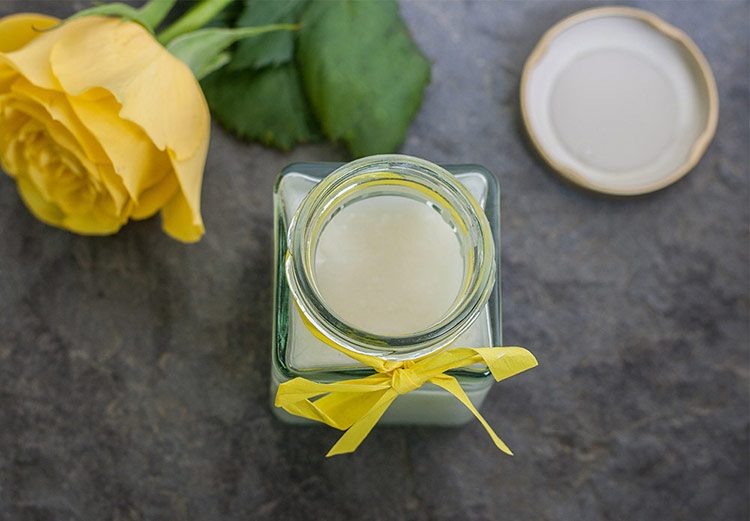 Draufsicht auf einen geöffneten Glas-Tiegel mit Creme-Deo, im Hintergrund eine gelbe Rose