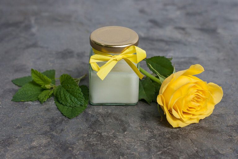 Creme-Deo in einem Glastiegel mit gelber Schleife, daneben eine gelbe Rose und Zitronenmelisse-Blätter